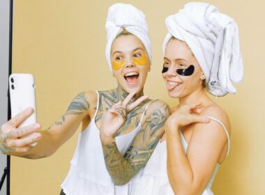 Two women enjoying a skincare routine with facial masks, taking a selfie wrapped in towels against a beige background.