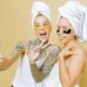 Two women enjoying a skincare routine with facial masks, taking a selfie wrapped in towels against a beige background.